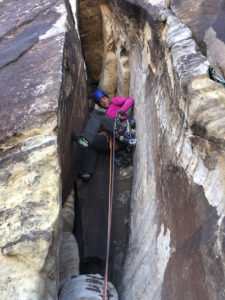 woman rock climbing in grey rock climbing pants and pink coat